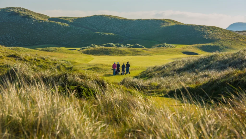 A group of golfers walking across a scenic green course surrounded by rolling hills and tall grass.