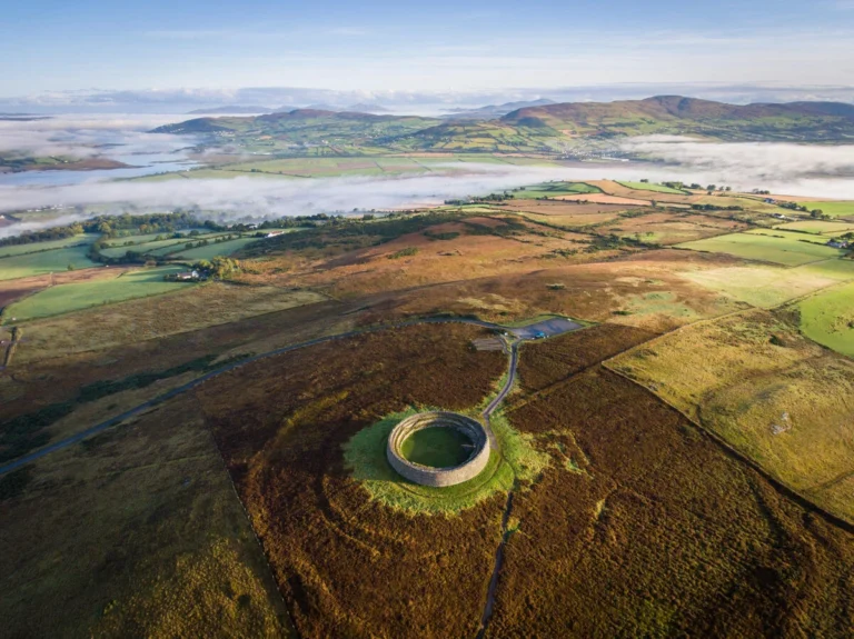 Aerial view of a historic stone fort surrounded by lush green fields and misty mountains in Ireland.