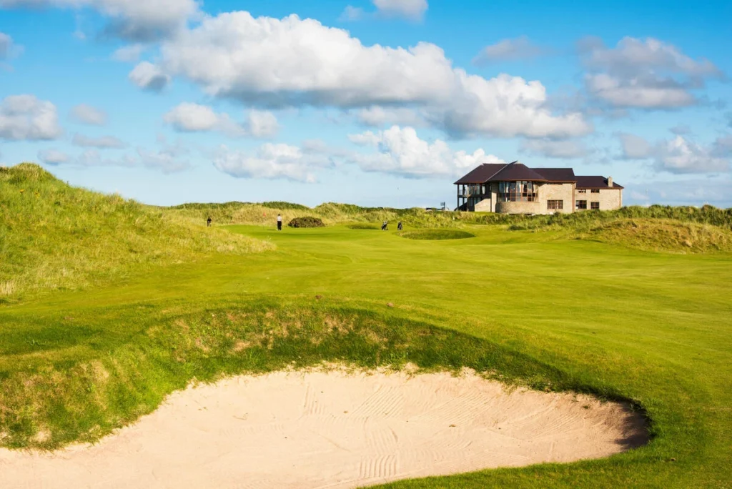 Scenic golf course with rolling greens, a sandy bunker, and a clubhouse under a bright blue sky. Ideal for golfers.
