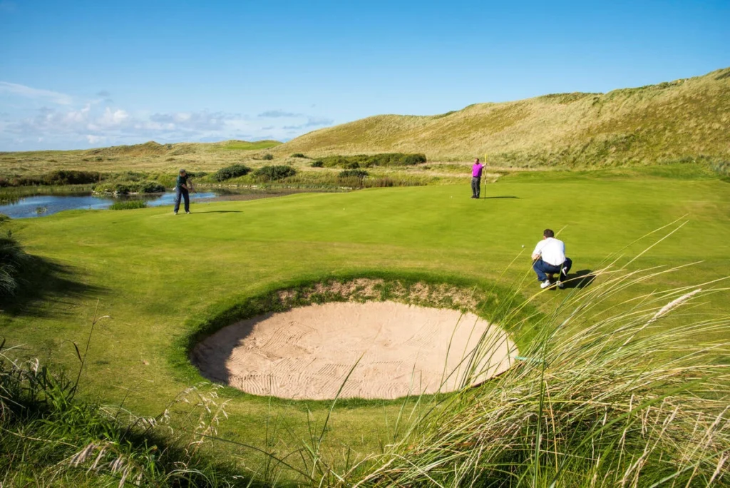 Golfers putting on a lush green course near a bunker, with scenic hills and a clear blue sky in the background.