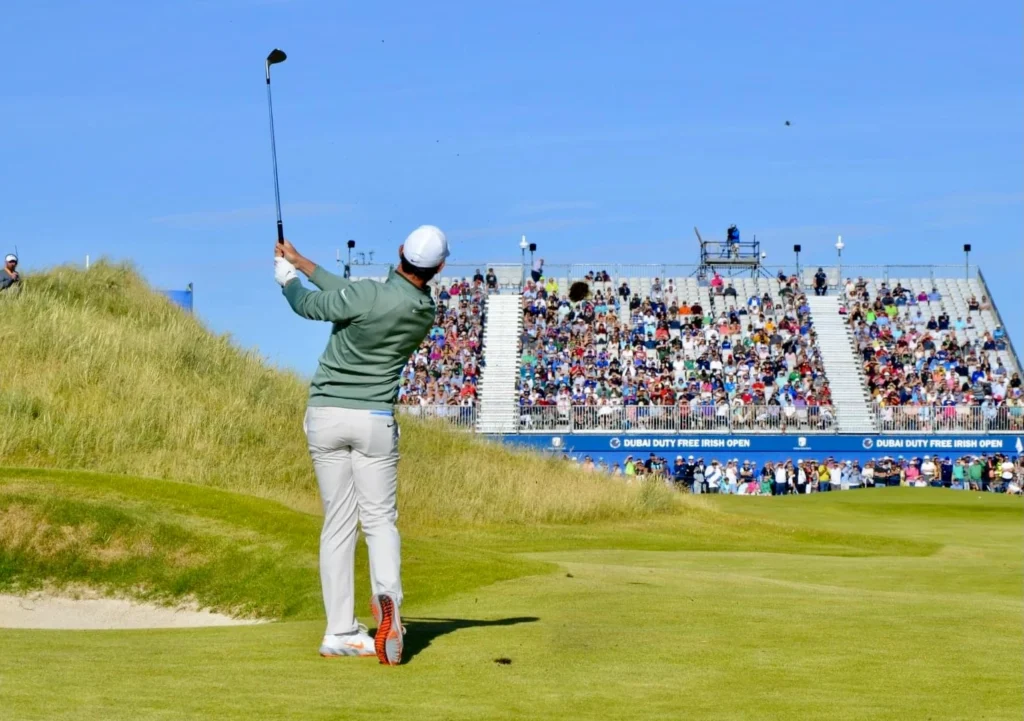Golfer hitting a shot at the Dubai Duty Free Irish Open, with a lively crowd and clear blue skies in the background.