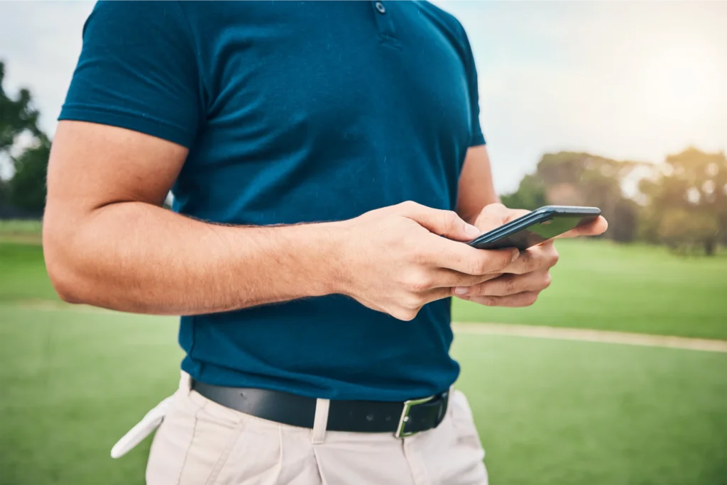 Man in a blue polo shirt using a smartphone on a golf course, enjoying outdoor leisure time.