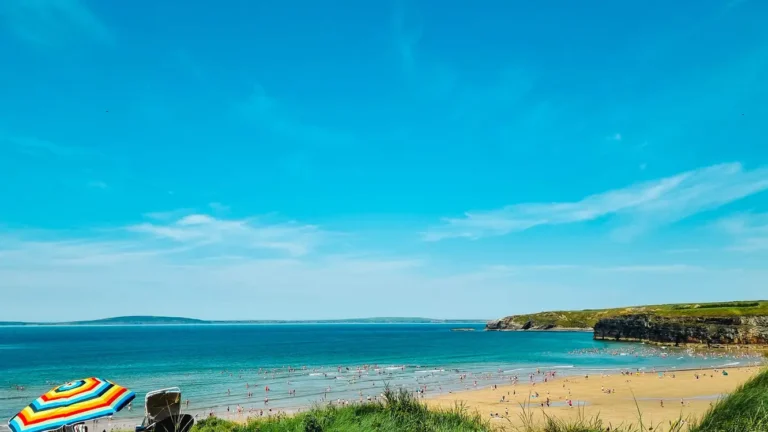 Busy Irish beach with swimmers, golden sand and cliffs, rainbow umbrella in the foreground.