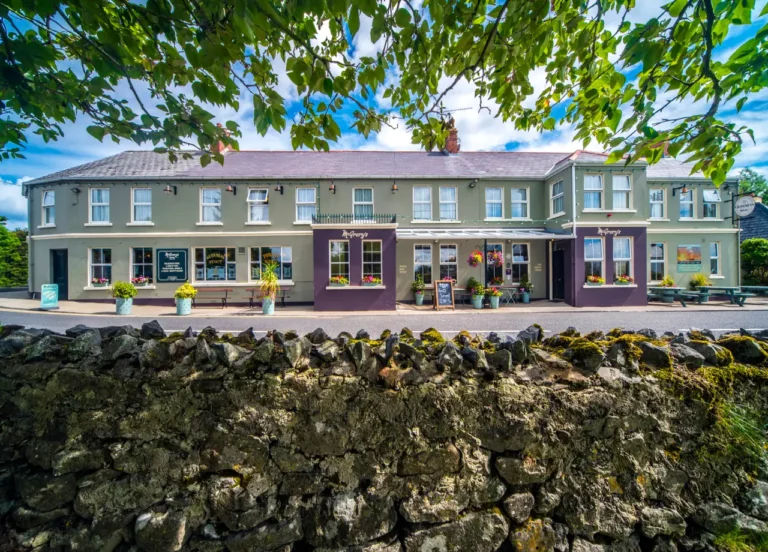 Charming pub with colorful flower pots, stone wall, and sunny skies; perfect for a cozy drink in Ireland.